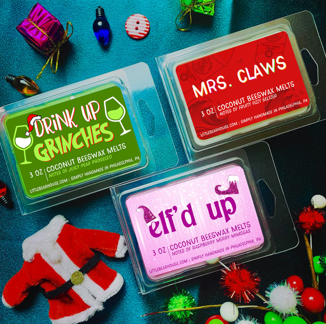 3 oz Drink Up Grinches, Mrs. Claws, and Elf'd Up beeswax and coconut wax melt on blue background with Christmas lights, gift boxes, and a Santa coat. Simply handmade in Philadelphia, PA,